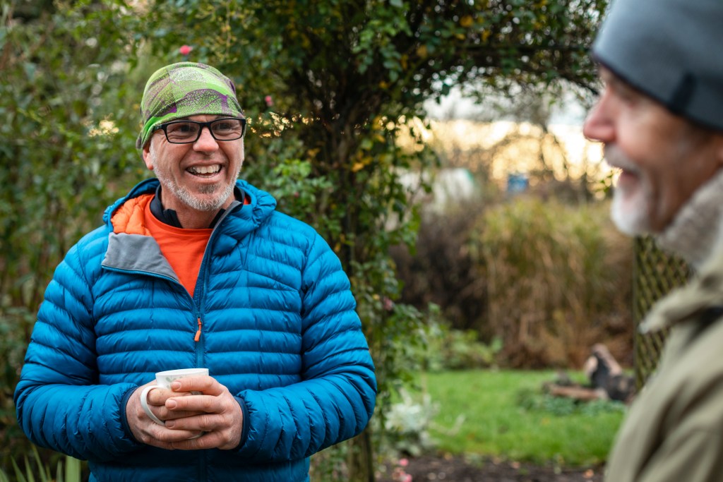 Volunteers sitting outdoors wearing warm casual clothing on a sunny cold winters day. They are resting and having a tea break. They are laughing and talking together, drinking hot drinks.