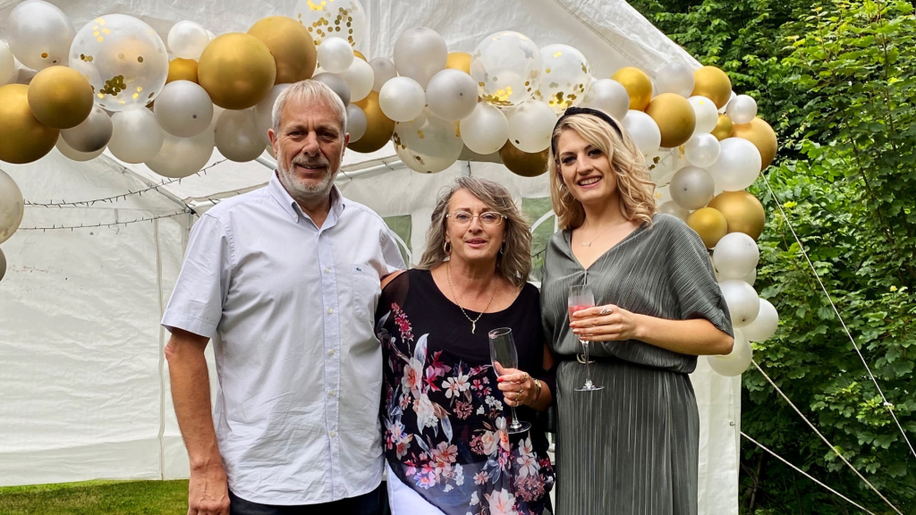 A photo of Sue, Shanice and Mike stood in a garden at a party. There are gold balloons and they are holding champagne flutes.