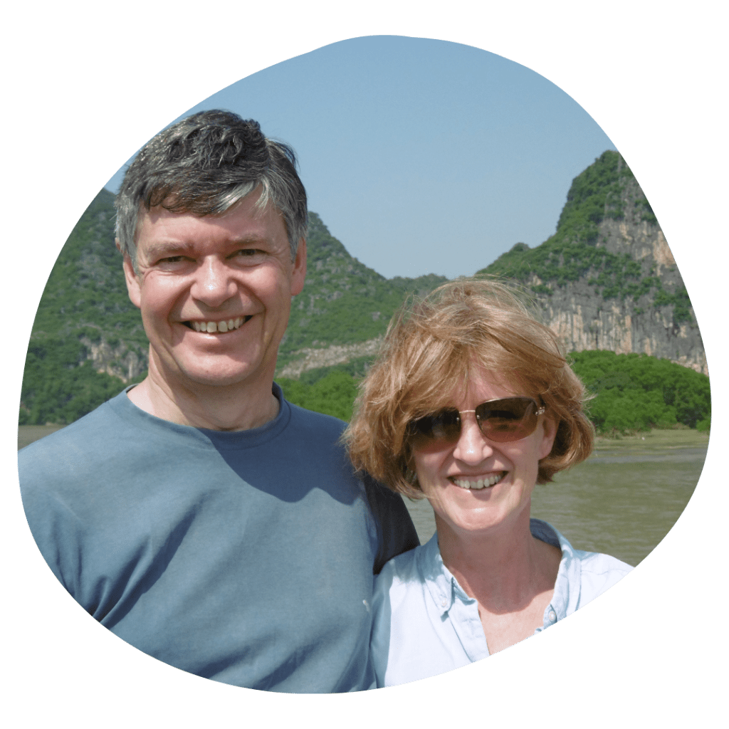 A photo of a man, short greyish hair wearing a blue-grey jumper, and woman, bobbed strawberry blonde hair wearing sunglasses, stood infront of mountains in a circle.