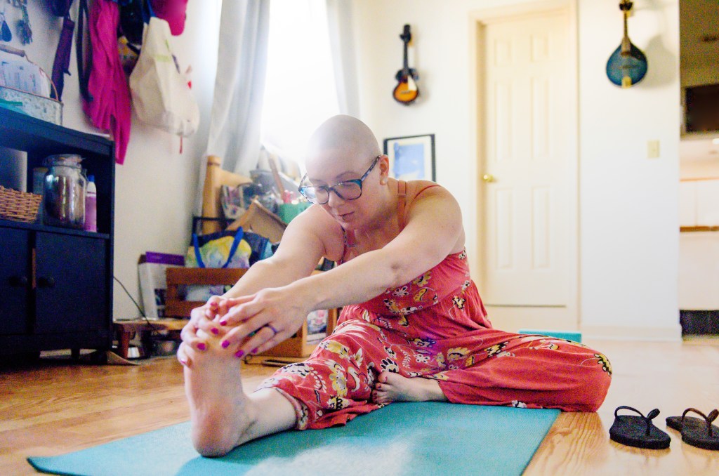 A Millennial woman in her 30s with breast cancer practices yoga in the morning at home. Her head is bald.