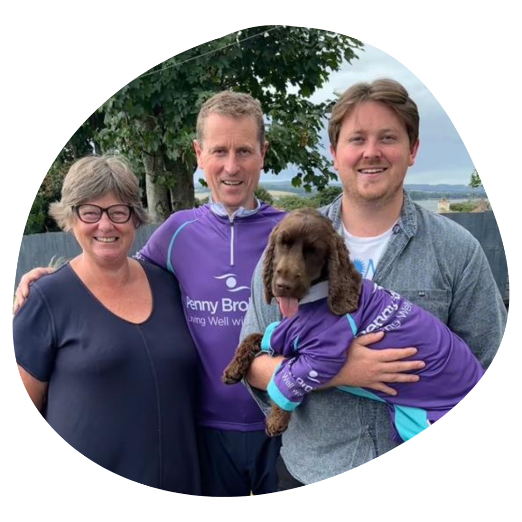 Photo of a family of three, left to right, woman with grey bobbed hair wearing glasses, man with short hair wearing a cycling jersey and shorts, young man wearing a blue/grey shirt over a white tshirt and shorts, holding brown curly haired dog wearing a cycling jersey.