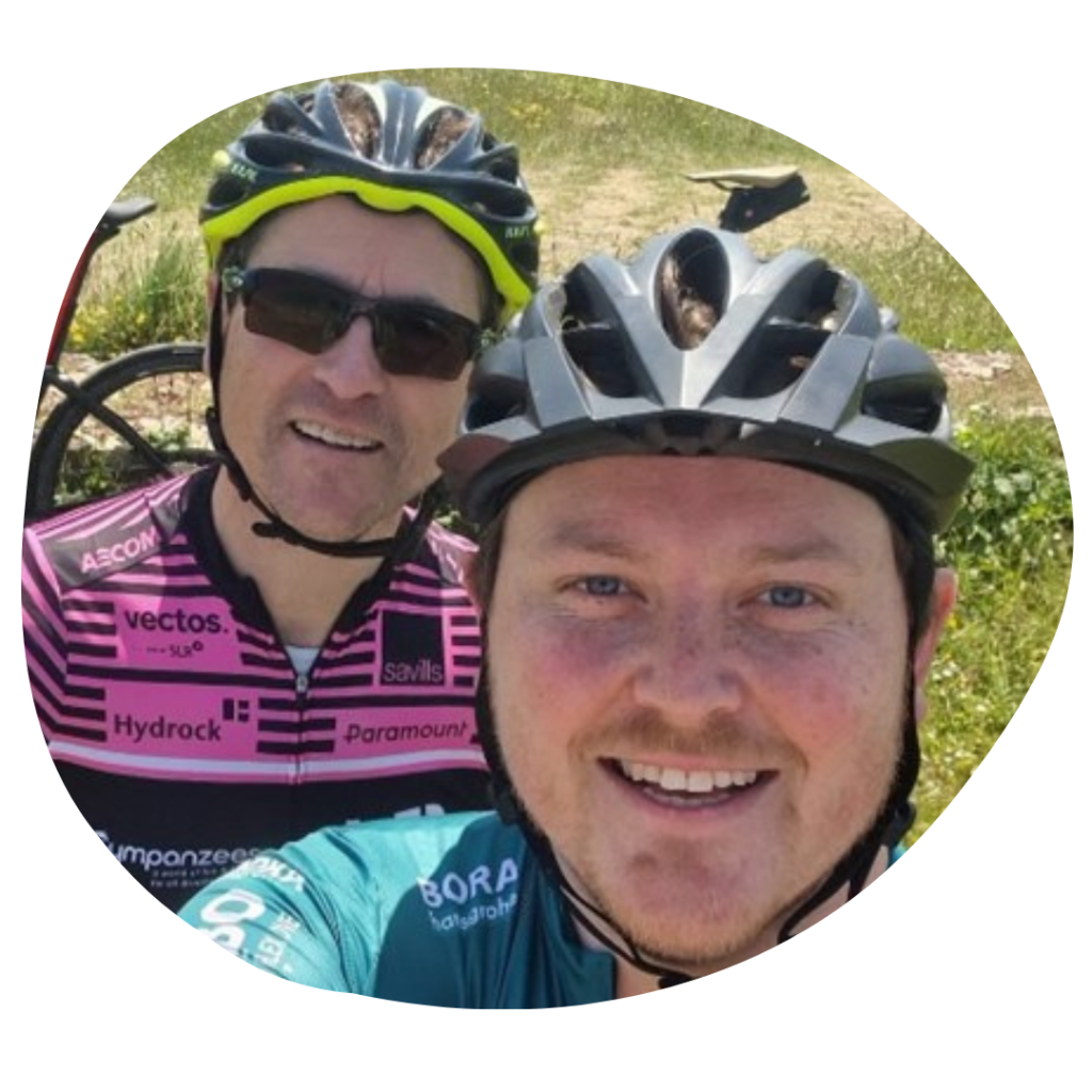 Two men, left to right, one wearing a pink cycling jersey, sunglasses and a bike helmet with yellow trim, the other wearing a teal-blue cycling jersey, smiling with a black helmet.